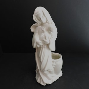 May include: A white ceramic figurine of a woman holding a child, with a vase-like structure at the base. The woman is draped in flowing robes and a head covering. The figurine is a religious or decorative item.