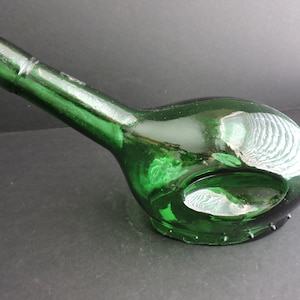 Puede incluir: Una botella de vidrio verde con un cuello largo y un cuerpo bulboso. La botella tiene una forma única con una gran abertura en el lateral. El vidrio tiene una ligera textura y es de un color verde intenso.
