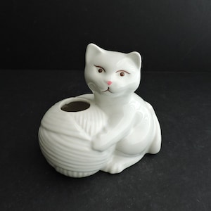 Puede incluir: Figura de gato de cerámica blanca sosteniendo un ovillo de lana. El gato tiene detalles rosas en la nariz y los ojos. La figura tiene un agujero en la parte superior, posiblemente para una pequeña planta o decoración. El gato está sobre una superficie negra.