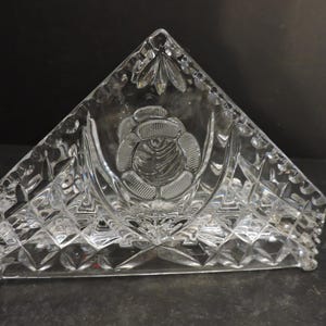 Peut inclure: Porte-serviettes en verre transparent de forme triangulaire. Il présente un motif de rose à l'avant et un motif de losanges sur les côtés. La base a de petits pieds. Un article décoratif pour la table.
