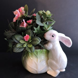 May include: A white ceramic bunny figurine leans against a green and white cabbage-shaped planter filled with pink and green flowers. The bunny has pink inner ears and a sweet expression, creating a charming Easter or spring decoration.