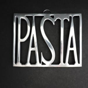May include: A silver-coloured metal wall hanging in a rectangular shape. The word "PASTA" is cut out of the metal in a bold, sans-serif font. A small loop is at the top for hanging. The background is black.