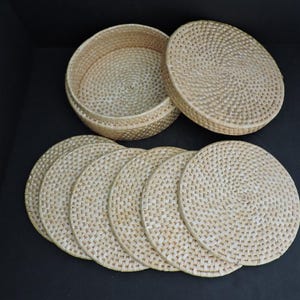 May include: A collection of woven rattan coasters and a matching round container with a lid. The coasters and container are light beige and feature a textured, woven design. The container is open, showing its inside.