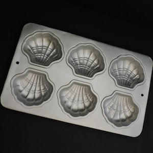 May include: A silver madeleine tin featuring six shell-shaped moulds. The rectangular tin has rounded corners and small holes. The moulds are detailed with shell patterns, ideal for baking madeleine cakes. The pan measures approximately 25 cm in length.
