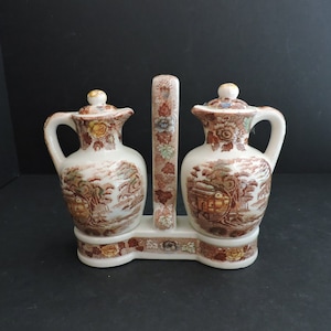 May include: A ceramic oil and vinegar set with a cream-colored base and brown transferware landscape design. The set includes two small pitchers with handles and lids, connected by a central handle.