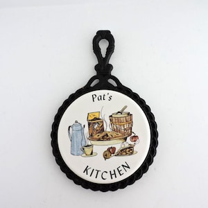 Puede incluir: Un salvamanteles redondo con marco de hierro fundido negro y un azulejo de cerámica central. El azulejo presenta una ilustración colorida de una cafetera azul, un pastel y un cubo de madera, con el texto Pat's KITCHEN en negro.