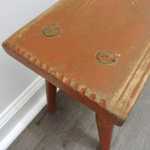 May include: A small, antique wooden stool with a rectangular seat and three legs. The stool is painted in a distressed orange color with visible wear and tear, and measures approximately 12 inches long.