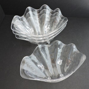 May include: Set of clear, clam-shaped serving bowls. The bowls have a wavy, scalloped edge and a textured interior. The bowls are made of a transparent material, and are stacked in a group of four, with two bowls in the foreground.