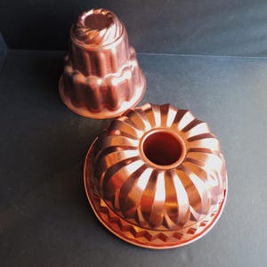 May include: Two copper bundt cake pans. One pan is a traditional shape with a fluted design, and the other is a smaller, tiered design. Both pans have a shiny, reflective surface and are set against a dark background.