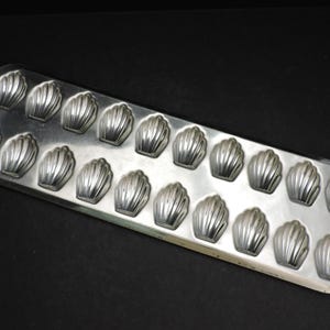 May include: A rectangular, silver-colored madeleine pan with 16 shell-shaped molds. The pan is made of metal and sits on a black surface. The molds are evenly spaced and have a ribbed design.