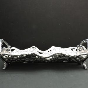 May include: A rectangular silver-toned metal tray featuring an intricate, raised floral and vine relief pattern across its sides. The tray has two ornate handles on the ends and is supported by four small, decorative feet, creating a raised base.