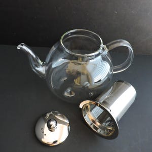 May include: A clear glass teapot with a curved handle and spout, accompanied by a removable cylindrical stainless steel mesh infuser and a polished silver-tone metal lid featuring a small knob and three steam vent holes near the edge.