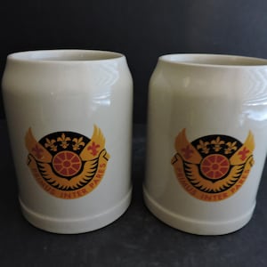 Può includere: Due boccali da birra in ceramica bianco sporco. Ogni boccale presenta uno stemma con un design nero, rosso e giallo, incluso il testo "PRIMUS INTER PARES". I boccali hanno una forma cilindrica e una parte superiore leggermente svasata.