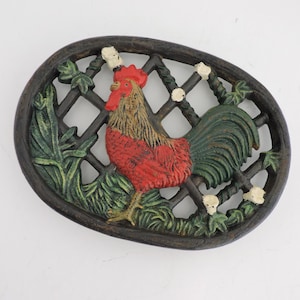 May include: Oval cast iron rooster wall decor. The rooster is painted red, green, and brown, set against a black lattice and foliage background. The decorative piece features a detailed design and is suitable for indoor or outdoor use.