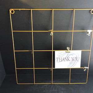 Puede incluir: Una rejilla de alambre dorada con forma rectangular, con un patrón de cuadrícula. Pequeños clips están unidos a la rejilla. Una tarjeta blanca con las palabras "THANK YOU" está sujeta a la rejilla.
