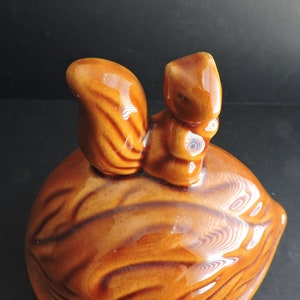 May include: A ceramic squirrel figurine perched atop a walnut-shaped container. The squirrel and walnut are glazed in shades of brown, with detailed textures. The figurine is likely a decorative item or a container for small items.