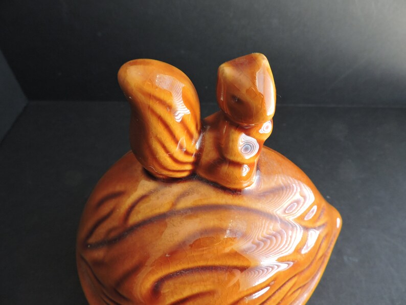 May include: A ceramic squirrel figurine perched atop a walnut-shaped container. The squirrel and walnut are glazed in shades of brown, with detailed textures. The figurine is likely a decorative item or a container for small items.