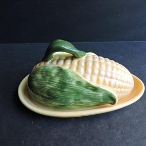 May include: A ceramic corn-shaped butter dish with a yellow base and a corn cob lid. The lid is detailed with a green husk and a green handle. The dish is designed to resemble a realistic ear of corn.