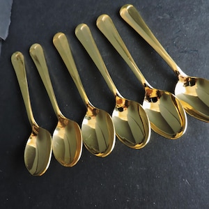 Puede incluir: Seis cucharas doradas dispuestas en diagonal sobre una superficie oscura. Las cucharas tienen un acabado brillante y reflectante, con mangos alargados y cuencos ovalados. Las cucharas son probablemente para servir o comer.