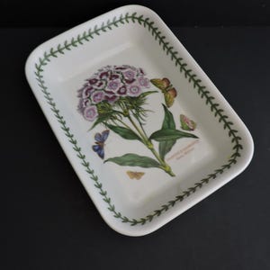 Puede incluir: Una fuente de horno rectangular de cerámica blanca con una ilustración botánica de una flor Dianthus barbatus morada y blanca con hojas verdes y varias mariposas pequeñas. El borde está decorado con un patrón de hojas verdes.