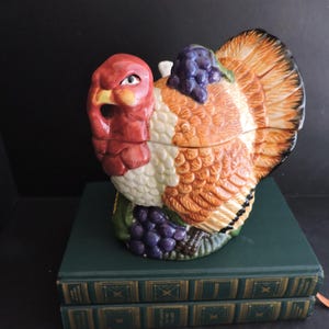 Op de afbeelding: Een keramische kalkoenvormige container met deksel, voorzien van een rode kop en nek, witte en oranje gestructureerde veren, en trossen paarse druiven aan de basis en op de rug, met een opvallende snavel en gedetailleerd verenkleed.