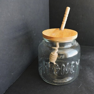 May include: A clear glass honey jar with the word "HONEY" embossed on the front. It has a wooden lid and a wooden honey dipper. The jar is on a dark surface.