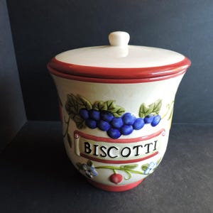 May include: A ceramic biscotti jar with a lid. The jar is off-white with a red rim and base. It features a design of blue grapes, green leaves, and the word "BISCOTTI" in a banner.