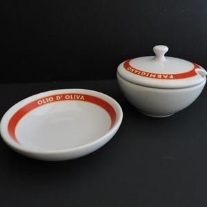 May include: A white ceramic bowl and a lidded container with a matching orange band. The bowl features the text OLIO D' OLIVA in white, while the lidded container displays the text PARMIGIANO in white on the orange band and includes a small notch.