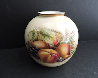 May include: A round, cream-colored vase with a painted fruit and leaf design. The artwork features peaches, grapes, and foliage in shades of red, green, and brown. The rim is trimmed with a gold band.