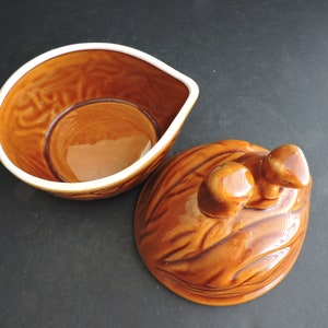 May include: A brown ceramic serving dish with a lid. The dish has a unique shape with a white rim and a textured surface. The lid is shaped like a walnut with two small knobs.