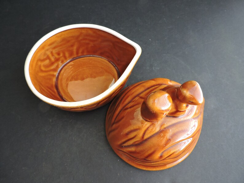May include: A brown ceramic serving dish with a lid. The dish has a unique shape with a white rim and a textured surface. The lid is shaped like a walnut with two small knobs.