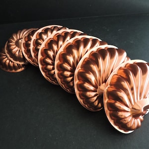 May include: A collection of copper molds, each with a fluted, shell-like design. The molds are arranged in a row, showcasing their intricate details and warm, metallic color. The kitchenware is displayed on a dark surface.