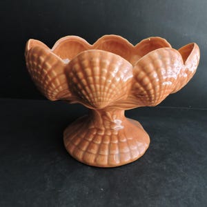 May include: A ceramic pedestal bowl in a warm, orange-brown hue. The bowl is shaped like a series of seashells, with a scalloped rim and a textured surface. The base is also shell-like, providing a stable foundation for the decorative piece.