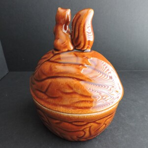 May include: A brown ceramic nut-shaped container with a lid, featuring two squirrel figurines on top. The container has a glossy finish and is designed to resemble a walnut. The squirrels are facing each other.