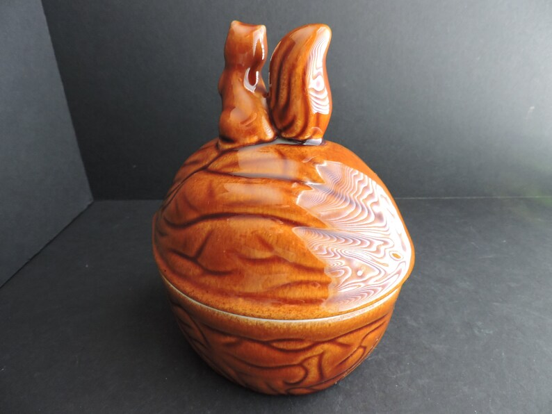 May include: A brown ceramic nut-shaped container with a lid, featuring two squirrel figurines on top. The container has a glossy finish and is designed to resemble a walnut. The squirrels are facing each other.