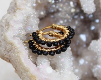 Pode incluir: Brincos de argola dourados com contas pretas. As argolas são enroladas com fio dourado e apresentam uma fileira de pequenas contas pretas redondas. Os brincos são exibidos em uma superfície branca texturizada.