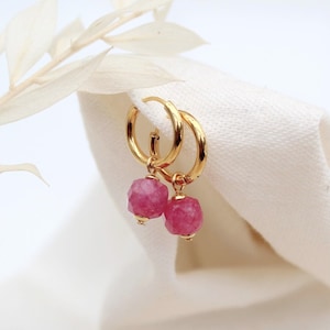 May include: Gold hoop earrings featuring faceted, pink gemstone drops. The earrings are presented on a white cloth, with a sprig of dried flowers in the background. The earrings have a simple, elegant design.