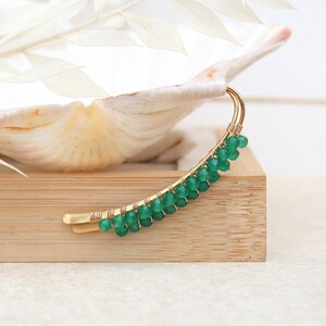 Op de afbeelding: Een gouden draad oorcuff met groene edelsteen kralen. De cuff is gebogen en heeft een delicaat ontwerp. De oorcuff wordt getoond op een houten oppervlak.