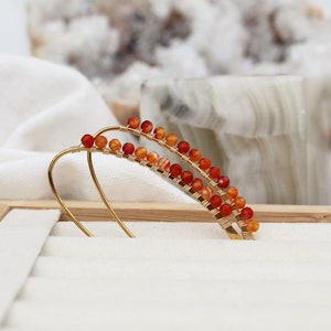 Könnte beinhalten: Goldfarbene Ohrkletterer mit Reihen aus kleinen, orange-roten Perlen. Die Perlen sind auf dünnem Golddraht aufgefädelt und bilden ein gebogenes Design. Die Ohrringe werden auf einer weißen Oberfläche mit einem Holzrahmen im Hintergrund präsentiert.
