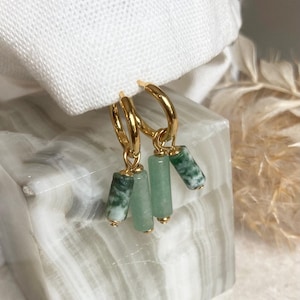 May include: Gold hoop earrings featuring three hanging green and white cylindrical beads. The earrings are presented on a white surface, with a soft, neutral background. A fashionable accessory.