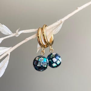 May include: Gold hoop earrings featuring two spherical pendants with a mosaic design. The spheres are dark blue with iridescent, square inlays. The earrings hang from a gold hoop and are set against a neutral backdrop.