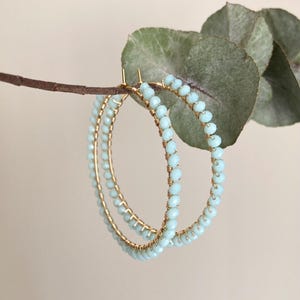 Può includere: Un paio di orecchini a cerchio dorati adornati con perline azzurre. Le perline sono infilate lungo il filo d'oro, creando un accessorio delicato ed elegante. Gli orecchini sono esposti su uno sfondo neutro con un rametto di verde.