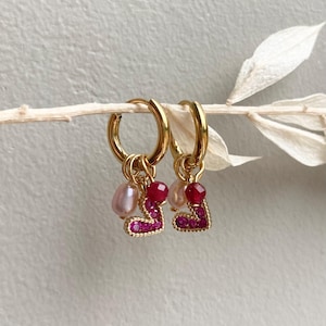 May include: A pair of gold hoop earrings with heart-shaped charms. Each earring features a small pink pearl, a red gemstone, and a heart outlined with small pink gemstones. The earrings are displayed on a light-colored twig.