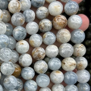 May include: A collection of round, polished aquamarine gemstone beads. The beads display a range of pale blue, white, and tan colors, with variations in translucency. The beads are smooth and spherical, suitable for jewelry making.