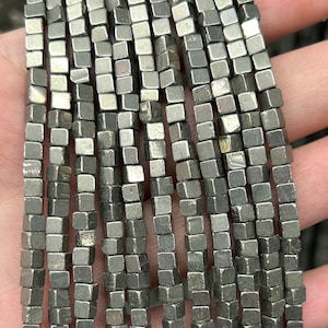 Può includere: Una collana di perline quadrate color argento. Le perline sono in pirite e hanno una texture leggermente ruvida.
