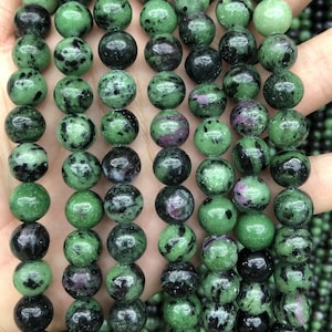 May include: A strand of green, black, and pink gemstone beads. The beads are round and have a speckled appearance.