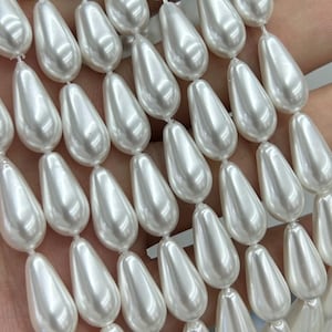 May include: A collection of white, teardrop-shaped faux pearl beads arranged in vertical rows. Each bead features a smooth, glossy finish with a reflective surface, strung together to create a uniform pattern of elongated, pearlescent shapes.
