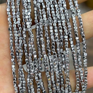 Può includere: Primo piano di più fili di piccole perline cubiche grigio metallizzato. Le perline sono infilate insieme, creando una superficie strutturata e riflettente. Le perline sono uniformi per dimensioni e forma, ideali per la creazione di gioielli o progetti artigianali.