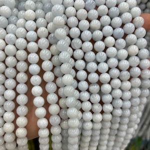 May include: Strands of round, pale blue-gray gemstone beads. The beads are uniform in size and have a smooth, polished surface. The beads are strung together, creating a dense, textured pattern. The overall effect is a soft, luminous appearance, suitable for jewelry making.