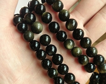 Könnte beinhalten: Eine Kette aus schwarzen Obsidianperlen mit goldenen Sprenkeln. Die Perlen sind rund und glatt und auf einer durchsichtigen Gummikordel aufgefädelt.
